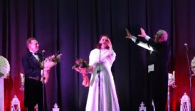 artyści na scenie, dwoje panów w garniturach, na środku kobieta w długiej białej sukience z białym kożuszkiem, wpatrzona w górę, w ręku trzyma bukirt kwiatów