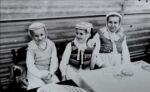Gminne dożynki na stadionie w Pępowie 15 września 1985 rok ph. ,,Otwórzcie szeroko wrota, bo się zakończyła żniwna robota". dwie dziewczynki i babcia w ludowych strojach siedzą przy stole i się uśmiechają
