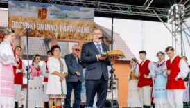na pierwszym planie mężczyzna trzyma chleb na tacy i przemawia przez mikrofon na statywie, w tle osoby ubrane w stroje ludowe i baner gminno-parafialne