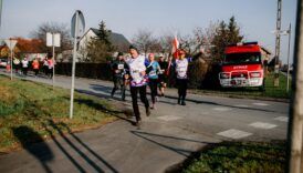 biegacze z numerkami startowymi i biało-czerwoną flagą. w tle wóz strażacki